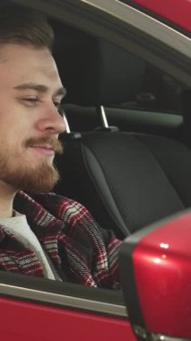 Güzel bakımlı sakallı şık bir genç adam sürücünün koltuğundan neşe saçan göz alıcı kırmızı arabasıyla kendine güveni ve memnuniyeti yayıyor. Rahat duruşu ve büyüleyici gülüşü yeni sahipliğin heyecanını yansıtıyor.