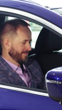 Sivri bir ekose takım elbise giymiş şık sakallı, bakımlı bir adam, parlak mavi bir arabanın sürücü koltuğunda güven içinde oturur. Çekici gülüşü profesyonelliği yansıtıyor. Şatafatlı iç güzelliği inceliyor, coşkusunu gösteriyor.