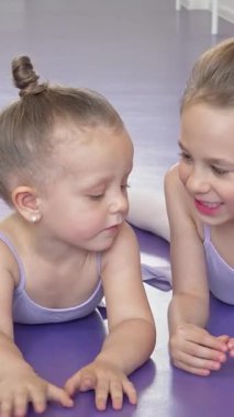 Pastel tayt giymiş, canlı mor bir zeminde dinlenen, sırlarını fısıldayan ve peluş oyuncaklarıyla kıkırdayan iki sevimli balerin. Bu hoş sahne çocukluk arkadaşlığının ve dans etmenin güzelliğini özetliyor.