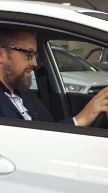 Şık, olgun bir beyaz adam, galeride el değmemiş beyaz bir aracın vitrininden arabanın anahtarını gururla tutarken mutluluk saçıyor. Bakımlı sakalı ve şık kıyafeti bu gururlu anın heyecanını arttırıyor.