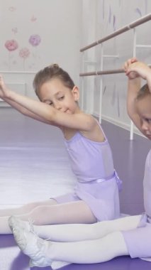 İki küçük sevimli balerin, parlak dans stüdyosunun canlı mor zeminiyle güzel bir tezat oluşturan yumuşak lavanta taytları bir arada geriniyor. Bu hoş sahne çocukluk sevincini ve balenin adanmışlığını yansıtıyor.