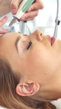 Huzurlu genç bir kadın, alnında ultrasonik bir fırçayla süzülen yetenekli bir güzellik uzmanının doğal parıltısını arttırması gibi rahat bir şekilde yatar. Bu lüks yüz bakımı derin temizlik ve canlandırma vaad ediyor. Cildini pürüzsüz bırakıyor.