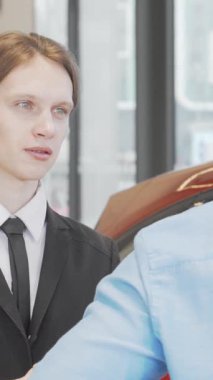 Çekici bir gülümsemesi olan şık genç bir satıcı, yeni olasılıkların heyecanını yansıtan parlak araçlarla çevrili lüks, modern bir araba galerisinde müşteriyle aktif olarak ilişki kuruyor. Bu canlı sahne otomotiv heyecanını temsil ediyor.