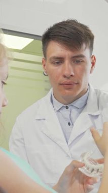 Şefkatli bir erkek dişçi, parlak ve modern bir klinikte meraklı bir genç kıza diş hijyeni teknikleri gösteriyor. Bu ilgi çekici sahne sıcaklığı ve profesyonelliği yansıtıyor, dişçilerin kendilerini sağlıklı beslenmeye adadıklarını vurguluyor.