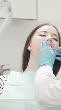 Neşeli genç bir kadın, dişçi koltuğuna yaslanıyor. Yetenekli bir erkek dişçi dişlerini dikkatlice incelerken ışıl ışıl gülüşü kendine olan güvenini yansıtıyor. Sakin renkler ve gelişmiş ekipmanlarla donatılmış şık, modern klinik güven verici bir şey yaratıyor.