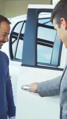 Modern bir araba galerisinde, bakımlı sakallı şık bir satıcı parıldayan beyaz bir aracın kapısını hevesle açar ve gülümseyen bir alıcıyı özelliklerini keşfetmesi için sıcak bir şekilde davet eder. Hava heyecan ve heyecanla dolu.