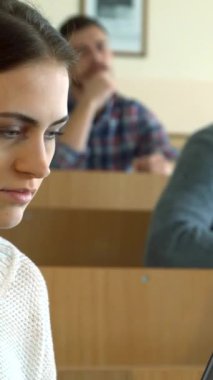 Dalgın bakışlı, meşgul, beyaz bir kız öğrenci parlak, geniş bir konferans salonunda, parlak dizüstü bilgisayarına özenle notlar yazar. Arka planda, ekose gömlekli iki erkek sınıf öğrencisi ve gri süveter giyen diğer erkek öğrencileri...