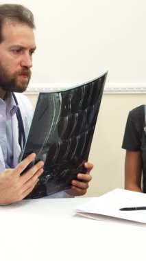 Güneşli bir ofiste, sıcak bir gülümsemesi olan genç bir erkek doktor özen ve profesyonellik hissi yayarak özenle reçete yazıyor. Dostane tavrı ve ışıl ışıl, davetkar atmosferi hastanın sağlığına olan bağlılığını yansıtıyor.