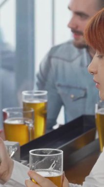 Canlı kızıl saçlı genç bir kadın, hareketli bir barda akıllı telefonuyla takılırken soğuk biranın tadını çıkarıyor. Canlı atmosfer kahkahalarla ve hareketli konuşmalarla uğulduyor, dostlar bir araya gelip, sosyal etkileşimin neşesini harmanlıyor.