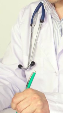 Beyaz önlük giymiş, odaklanmış beyaz bir erkek doktor titizlikle bir panoya parlak yeşil kalemle yazı yazıyor. Yumuşak, sıcak ışık sahneyi kaplıyor, özenli tavrını ve profesyonel atmosferini vurguluyor.