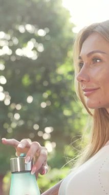 Canlılıkla parlayan ışıl ışıl bir kadın canlandırıcı bir açık hava egzersizinden sonra şişesinden ferahlatıcı bir yudum su içmekten zevk alıyor. Sıcak güneş ışığıyla yıkanmış ve canlı yeşilliklerle çevrili neşeli gülüşü sağlık ve neşe saçıyor.