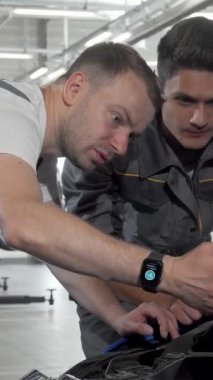 Gösterişli gri üniformalar içinde iki oto tamircisi, açık bir arabanın kaputuna dikkatle yaslanmış, motor arızası teşhisi konsantrasyonuyla dolu yüz ifadeleri. Modern garajdaki dinamik takım çalışması ve odaklanmış enerji.