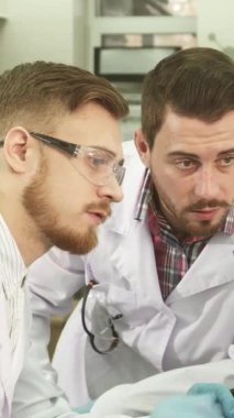 Çıtır beyaz önlükler ve koruyucu eldivenler giymiş iki genç erkek laboratuvar asistanı, bir bilgisayar ekranına dikkatle yaslanmış, test sonuçlarıyla ilgili ayrıntılı bir tartışmaya daldılar. İfadeleri merak ve kararlılık yayar.