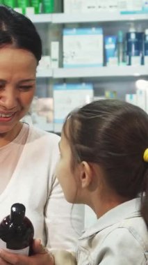 Neşeli bir eczanede renkli sağlık ürünleriyle dolup taşan neşeli bir anne meraklı kızıyla birlikte çareleri keşfederken keyifli bir anı paylaşıyor. Küçük kız, uzun siyah saçları arkadan bağlanmış ve gözleri ışıldıyor.