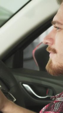 Neşeli, sakallı genç bir adam yepyeni arabasının gösterişli sürücü koltuğunda oturuyor ve kendine güvenen parlak araba anahtarlarını gösteriyor. Parlak gülüşü, sahipliğin heyecanını ve yeni maceraların heyecanını yansıtıyor.