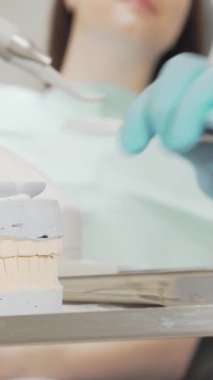 Bu yakından çekimde parlak metal bir tepside detaylı bir diş kalıbı sergilenirken, kendini işine adamış bir dişçi önlük ve eldiven giyerek arka planda tanınmayan bir hastayla ustalıkla ilgileniyor. İmaj profesyonellik ve özen gösteriyor.