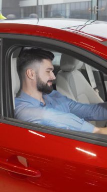 Zarif sakallı şık bir genç adam, galeride çarpan kırmızı bir arabanın sürücü koltuğunda güven içinde otururken mutluluk saçıyor. Bu canlı sahne, yeni bir araç seçmenin coşkulu anını özetliyor.