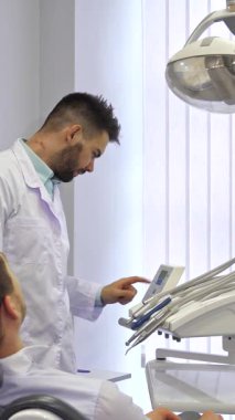 Kendini adamış beyaz önlüklü esmer bir erkek dişçi şık, modern bir diş sandalyesinde rahat bir şekilde oturan sarışın beyaz hastasıyla dikkatlice etkileşime girer. Parlak, havadar ofis, yumuşak doğal ışıkla aydınlatılır.