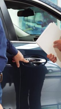 Modern bir araba galerisinde, yeni sahibi yeni arabasının sofistike kilitlerini heyecanla keşfeder. İki iyi giyimli adam, biri açık mavi gömlekli, diğeri şık ceketli, sıkı bir tokalaşmayı sembolize ediyor.