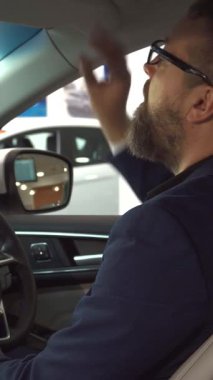 Şık sakallı, gözlüklü, gururlu bir adam galeride lüks bir aracın sürücü koltuğundan bir araba anahtarı tutuyor. Kendine güvenen gülüşü ve modaya uygun kıyafetleri davetkar bir ortam yaratırken modern iç mekan ve ferah