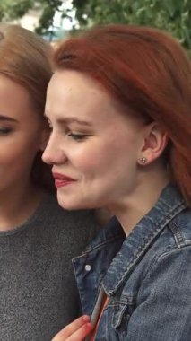 İki genç kadın, biri parlak kızıl saçlı, diğeri ise parlak sarışın, bir akıllı telefonu yemyeşil, güneşli bir parkta paylaşırken kahkahaya boğuluyorlar. Neşeli ifadeleri ve çevreleyen yeşillikler canlı bir atmosfer yaratıyor.