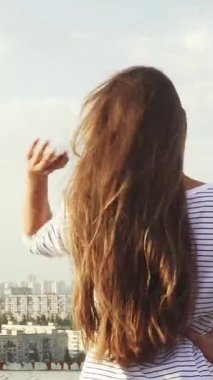 Genç bir kadın kameraya arkası dönük duruyor, uzun, dalgalı siyah saçları hafif meltemde sallanırken aşağıdaki çarpıcı şehir manzarasına hayran kalıyor. Sıcak akşam ışığı gökyüzünü altın rengi ile kaplıyor, canlı binaları vurguluyor ve...