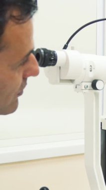 Odaklanmış genç bir adam, modern sağlık teknolojisinin doğruluğunu gösteren, parlak, steril bir ofiste, ileri teknoloji göz inceleme cihazına yaslanıyor. Özenli göz doktoru, görme sağlığının titizlikle yerinde olduğundan emin olmaya hazır.