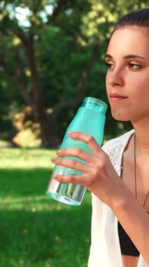 Huzurlu bir parkta, at kuyruklu koyu renk saçlı parlak genç bir kadın yoga seansları arasında ferahlatıcı sudan bir yudum alırken neşeyle ışıldıyor. Parlak yeşillik ve benekli güneş ışığıyla çevrelenmiş, onun mutluluk dolu ifadesi,