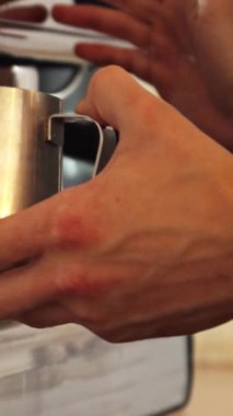 Rahat bir kafede, genç bir adam paslanmaz çelik sürahide ustalıkla süt buharı yapar, enfes bir kapuçino hazırlar. Odaklanmış elleri, duyarlılık ve sanatsallığı, taze demlenmiş kahvenin zengin aromasıyla dolu sıcak bir atmosfer yaratıyor.