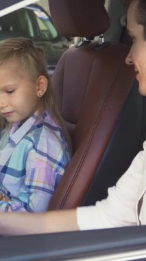 Renkli bir ekose gömlek giymiş keyifli küçük bir kız, yeni ve şık bir arabanın direksiyonunu sıkıca kavrıyor, babası yanında gururla parıldıyor. Zengin kahverengi deri ve modern özelliklerle süslenmiş lüks iç mekan,