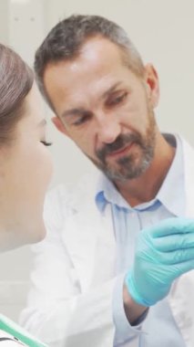 Neşeli bir kadın, dişçi koltuğunda otururken kendine güveni yayar. Parlak gülümsemesi kamerayı büyüler. Gıcır gıcır önlük ve eldiven giymiş özenli dişçi, gururla bir dişçi modeli sergiliyor.