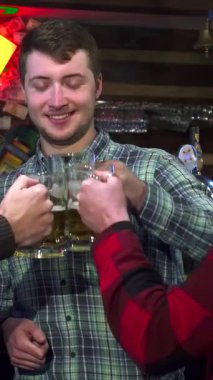 Üç neşeli arkadaş, şık damalı gömlekler içinde, kahkaha ve sıcaklıkla dolu canlı bir barda buzlu altın bira bardaklarını tokuşturuyorlar. Renkli ışıklarla süslenmiş köy barı, kutlayan davetkar bir atmosfer yaratıyor.