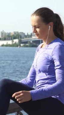 Kararlı genç beyaz bir kadın, ışıldayan bir nehirde bacaklarını açar ve enerji egzersizi için hazırlanır. Parlak at kuyruğu ve kulaklıklarıyla uzun siyah saçları ile, düşünceli bir şekilde şehrin ufuk çizgisine bakıyor.