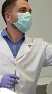 Kıtır beyaz önlüklü çalışkan bir orta yaşlı beyaz dişçi, klinik alanı sıcak bir ışıltıyla aydınlatarak parlak diş ışığını yakar. Bu yakın plan çekimde dikkatli esmer doktor, bir prosedür maskesi takıyor.