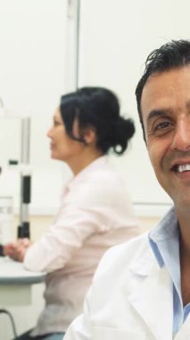 Neşeli bir doktor, şık mavi gözlüklü, kendine güveni tam, pozitif ışıldıyor. Arka planda, kendini işine adamış bir kadın doktor hastanın gözlerini dikkatle inceleyerek sıcak ve profesyonel bir ortam yaratıyor.