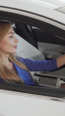 Neşeli bir kadın sürücü özgüven yayar, galerideki şık, modern bir arabanın sürücü koltuğundan başparmağını kaldırır. Canlı gülüşü ve şık kıyafetleri rüya aracını güvenceye almanın verdiği heyecanı yansıtıyor. Zarafeti somutlaştırıyor.