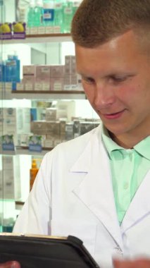 Gıcır gıcır beyaz bir tıbbi elbise giymiş odaklanmış bir erkek eczacı, tablet üzerinde gezinirken bir telefon konuşmasını ustalıkla yapıyor ve hasta bakımına olan bağlılığını gösteriyor. Düzenli ve canlı bir dizi ilaç ürünüyle çevrelenmiş bir şekilde,