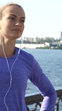 Genç beyaz bir kadın, kendine güvenen bir gülümseme ile kollarını uzatıyor, nehir manzarasının tadını çıkarırken etrafa bakıyor. Canlı bir menekşe bluzla giyinmiş, mp3 çalarıyla müzik dinliyor, ona hazırlanırken enerji ve coşku yayıyor.