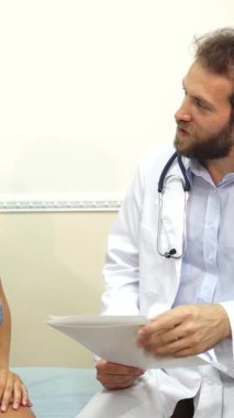 Parlak, profesyonel bir ofiste, kot giyen genç bir kadın, beyaz önlük giymiş doktoru olarak dikkatle dinliyor, ultrasonik araştırmasını tartışıyor. Atmosfer, tıbbi teçhizat ve yatıştırıcı ile vurgulanan odaklanma ve özenle doldurulmuştur.