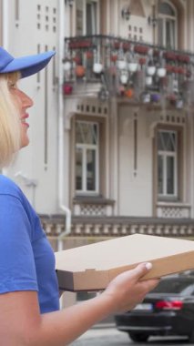 Parlak mavi üniformalı neşeli bir teslimatçı kadın dışarıda elinde pizza kutusuyla bir sonraki teslimatı için caddeyi tarıyor. Bu canlı sahne, onun bağlılığını ve istekliliğini vurgulayan fast food servisinin heyecanını yakalıyor.