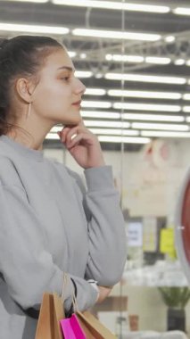 Atkuyruklu şık genç bir kadın, renkli bir mağaza vitrininden büyülenerek düşünceli bir şekilde duruyor. Alışveriş torbaları elinde hafifçe sallanırken, heyecan ve merak yayıyor, bu hareketli ortamda perakende keşfinin heyecanını somutlaştırıyor.