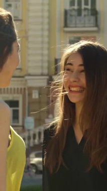 İki şık genç kadın, sıcak güneşin altında neşeli bir sohbet ederken, bir tanesi omzunda bir alışveriş torbasını dinlendiriyordu. Parlak gülümsemeleri ve büyüleyici mimari ile dolu canlı şehir arkaplanı bir atmosfer yaratıyor.