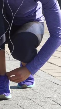 Kararlı genç bir kadın güneşli bir nehir yolunda çömeliyor, titizlikle göz alıcı mor spor ayakkabılarının bağcıklarını bağlıyor. Odaklanmış bakışları ve atletik kıyafetleriyle canlandırıcı bir açık hava macerasına hazırlanıyor. Enerji ve stili somutlaştırıyor.