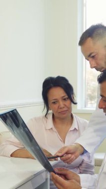 Parlak, modern bir muayenehanede genç bir kadın, canlı beyaz önlük giymiş iki doktor gibi dikkatle dinliyor ve röntgen sonuçlarını analiz ediyor. Oda profesyonellik ve empati yayarak hayati sağlık sezgilerinin olduğu destekleyici bir ortam yaratıyor.