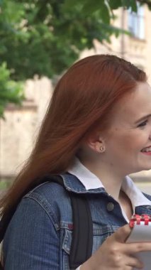 Canlı kızıl saçlı bir kız öğrenci yakışıklı erkek sınıf arkadaşıyla kampüste yemyeşil ve güneşli binalarla çevrili notlar hakkında konuşurken neşe saçıyor. Kahkahaları havada yankılanıyor, arkadaşlığın ve sevincin özünü yakalıyor.