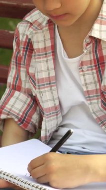 Canlı bir ekose gömlek giymiş sevimli küçük bir çocuk, yemyeşil bir çimenlikte defterine çizim yapmaya dalmış. Güneş ışığı ağaçların arasından süzülüyor, neşeli yüzüne sıcak bir parıltı saçıyor, kaygısız yaratıcılığın özünü yakalıyor.