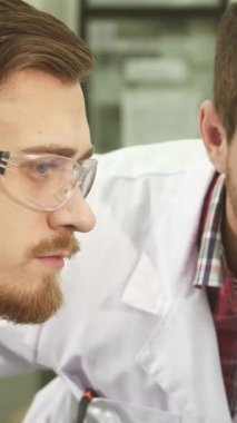 Yakın plan çekimde, analizlerinin ağırlığını yansıtan ciddi bir tartışmaya dalmış beyaz önlüklü iki genç erkek laboratuvar teknisyeni ortaya çıkıyor. Pürüzsüz, yüksek teknolojili ekipmanlarla çevrili...