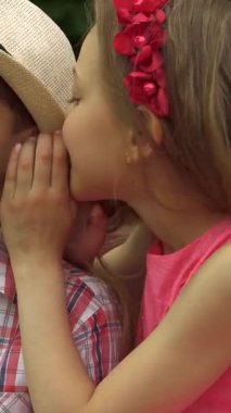 Küçük tatlı bir kız, güneşli bir bankta gülümseyen bir oğlanla bir sırrı paylaşmak için öne eğiliyor. Kahkahaları ılık yaz havasında yankılanıyor. Canlı yeşilliklerle çevrelenmiş, neşeli etkileşimleri çocukluğun saf neşesini yayıyor.