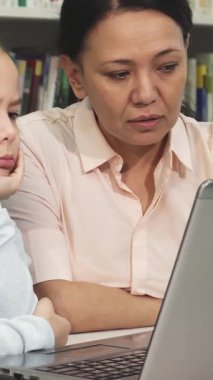 Canlı kitaplarla dolu samimi bir kütüphanede, yorgun küçük bir kız çenesini elinde dinlendirip dikkatle dizüstü bilgisayara bakıyor. Endişeli annesi eğiliyor, nazik rehberlik sunuyor, sevgi ve desteğin sıcaklığını somutlaştırıyor.