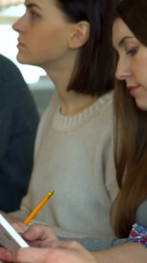 İki odaklanmış kız öğrenci, biri gösterişli kısa saçlı, diğeri uzun, dalgalı saçlı, canlı bir sınıf ortamında derinden bağlılar. Özenli ifadeleri ve şık kazakları modern eğitimin dinamik atmosferini yansıtıyor.